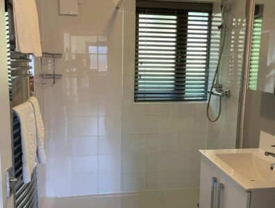 The Bathroom of Cottage 25 shows a spacious glass walled shower and a clean white sink. It has laminate wooden flooring and a metal rack for hanging towels and clothes.